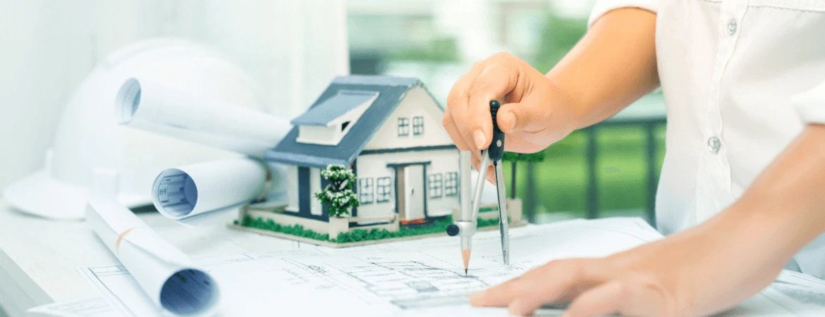 Architect designing house plans with blueprints and model home on desk.