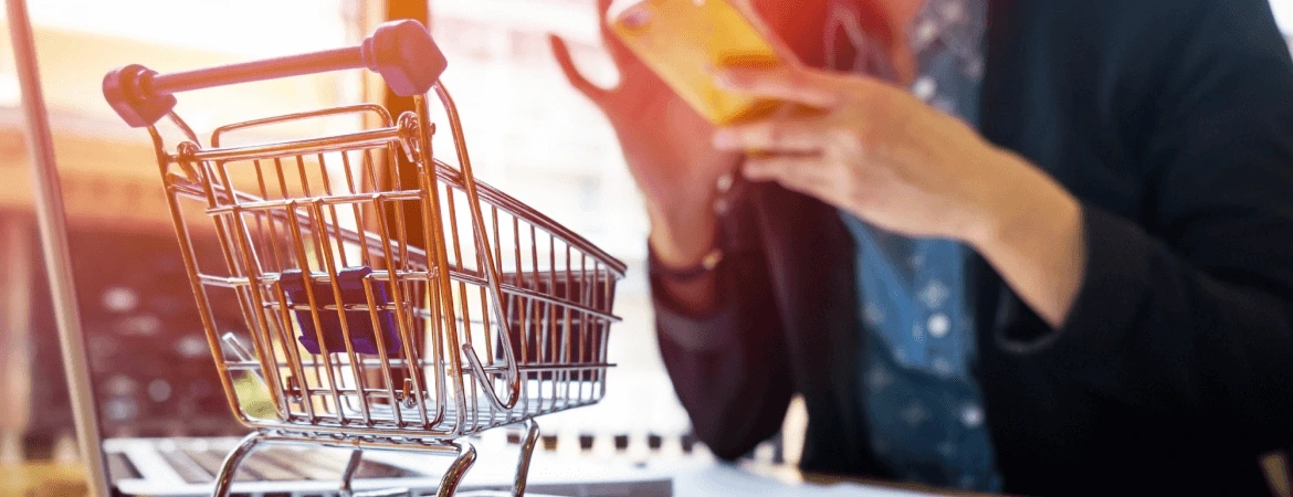 Shopping cart beside a laptop and a person using a smartphone for online shopping.